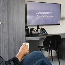 Hånd som holder en kaffekopp i forgrunnen, hotellrom med hotell-tv som infoskjerm i bakgrunnen.