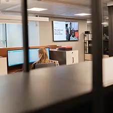 Kvinne som arbeider på en PC i et kontorlandskap. Infoskjerm på veggen.
