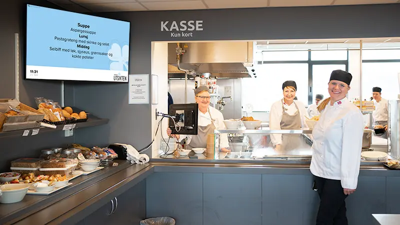 Kantine-personalet står ved siden av infoskjermen i kantina på Revmatismesykehuset. Oppslag med Dagens Meny vises på skjermen. Foto.